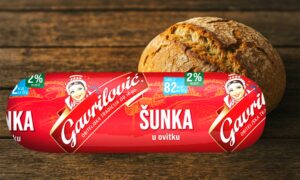 Gavrilović šunka u ovitku s oznakom 2% masti, ispred svježe pečenog kruha na drvenoj podlozi.