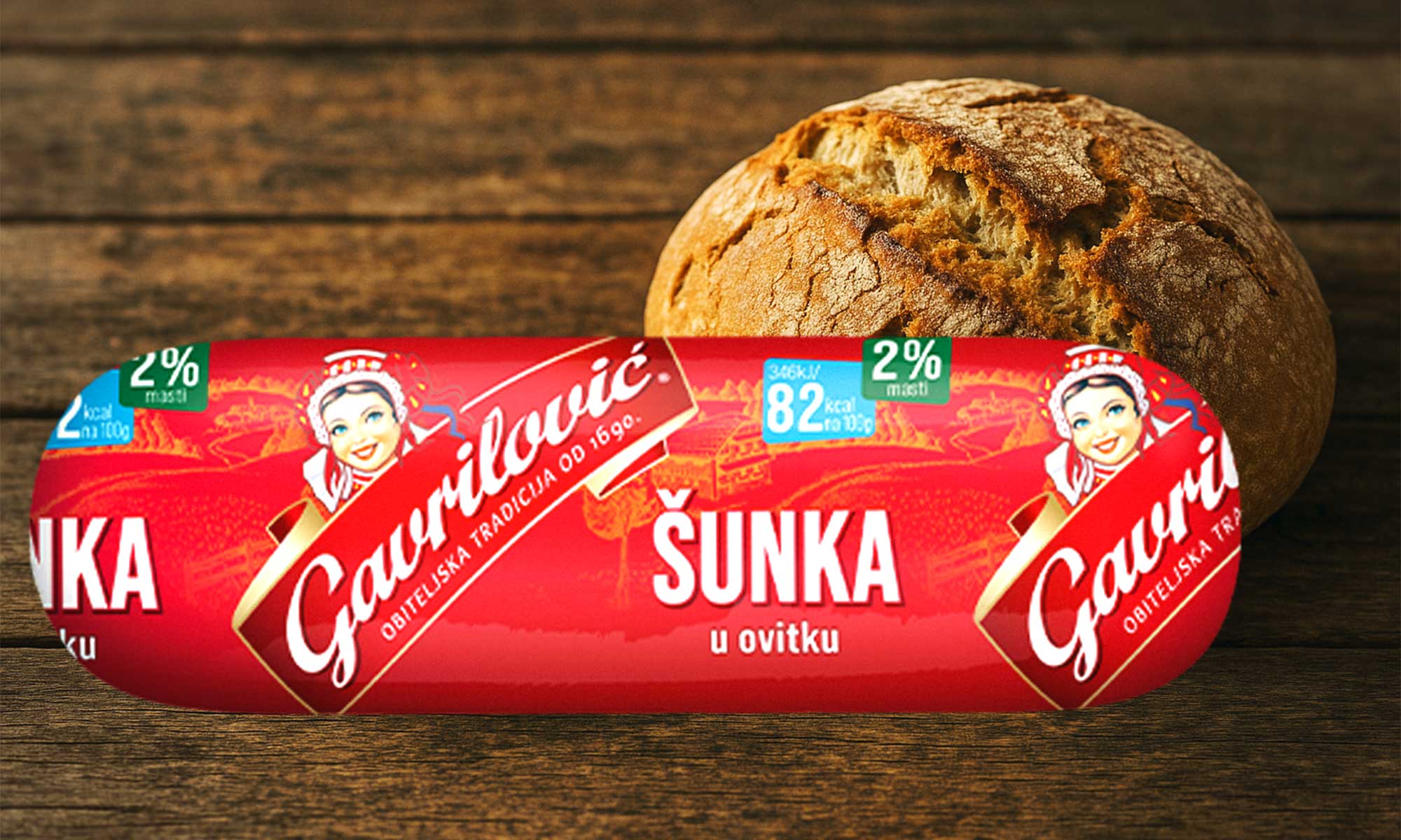 Gavrilović šunka u ovitku s oznakom 2% masti, ispred svježe pečenog kruha na drvenoj podlozi.