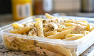 Plastična posuda s tjesteninom tipa penne i komadićima piletine, začinjena začinskim biljem, postavljena na kuhinjski pult u zatvorenom prostoru.
