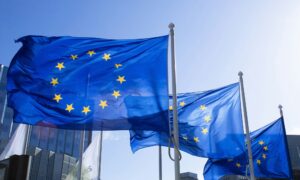 Tri europske zastave vijore se na vjetru ispred moderne poslovne zgrade, pod vedrim plavim nebom.